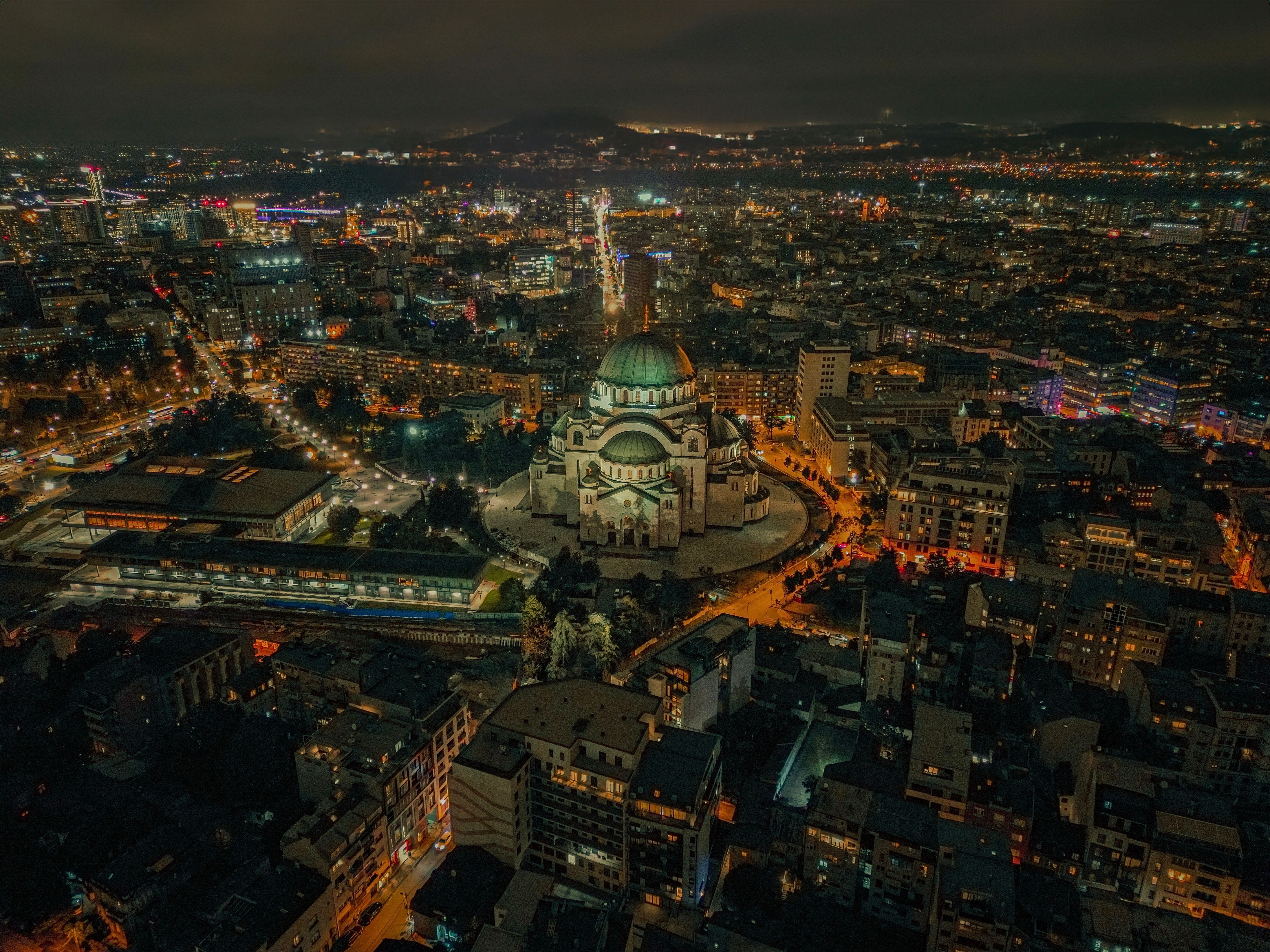 Flygfoto över Belgrad i kvällsljus med Hram Svetog Save i centrum och stadens ljus runt omkring.